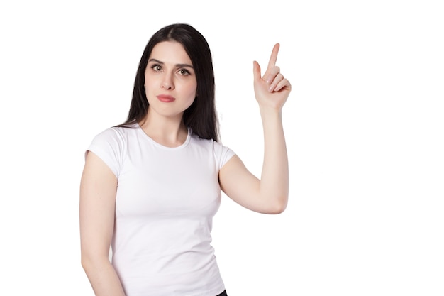 Joven atractiva mujer morena de pelo largo negro caucásico en camiseta blanca sobre fondo blanco, manos levantadas, dedo índice hacia arriba, idea de concepto de oferta comercial