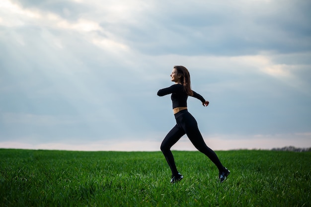 Joven atractiva mujer morena de pie en la naturaleza, vestida con leggings negros y un top negro. Día de verano, hierba verde. Cuerpo atlético y estilo de vida saludable.