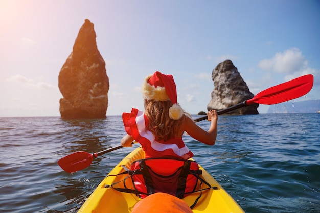 Joven atractiva mujer morena en traje de baño rojo y gorro de Papá Noel nadando en kayak alrededor de rocas de basalto