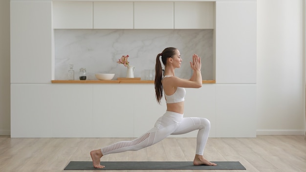 Joven y atractiva mujer multiétnica con pantalones blancos de ropa deportiva y top haciendo ejercicios de yoga de estiramiento en la estera de fitness en la sala de estar, salud y ejercicio, terapia mental en el concepto de hogar