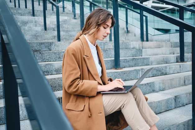 Joven y atractiva mujer de negocios casual con abrigo trabajando cuidadosamente en una laptop en las escaleras al aire libre