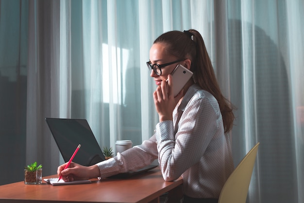 Joven y atractiva mujer de negocios con gafas está trabajando remotamente en casa utilizando un teléfono móvil y una computadora portátil.