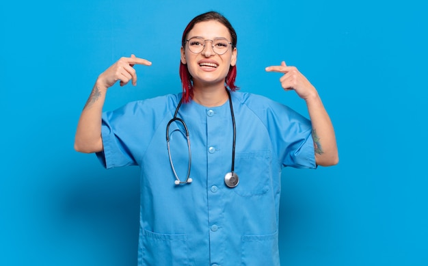 Joven atractiva mujer de pelo rojo sonriendo con confianza apuntando a poseer una amplia sonrisa, actitud positiva, relajada y satisfecha. concepto de enfermera del hospital