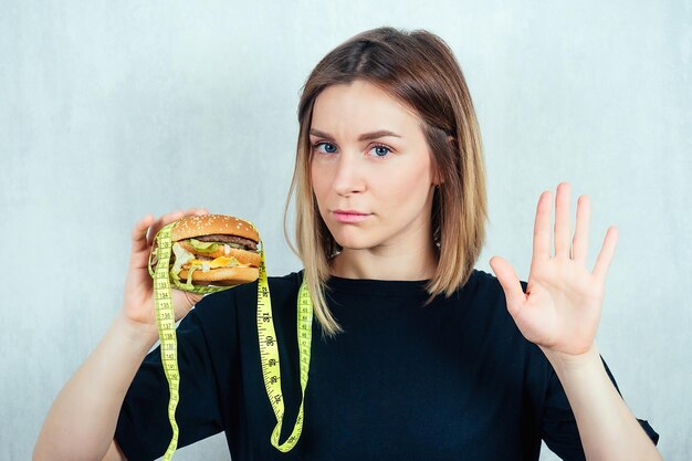 Joven y atractiva mujer rubia con camiseta negra y cinta métrica sostiene una hamburguesa alta en calorías y muestra un gesto de mano que detiene el concepto de rechazar la comida rápida y la dieta dañinas