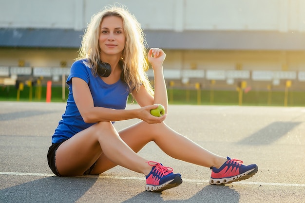 Joven y atractiva mujer rubia en ropa deportiva con auriculares sentado en el asfalto y comiendo una manzana verde en el estadio. estilo de vida deportivo y concepto de alimentación saludable