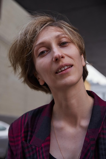 Joven atractiva mujer sonriente con el pelo corto con chaqueta roja a cuadros al aire libre frente al edificio. primer plano, retrato, de, caucásico, hembra