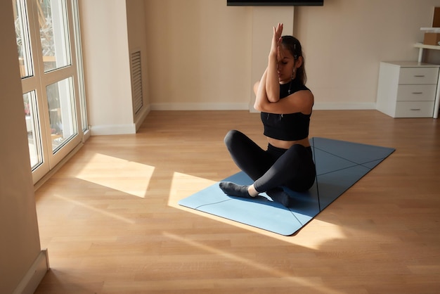 Joven atractiva mujer sonriente practicando yoga usando pantalones deportivos y top de cuerpo entero interior a