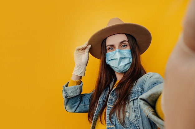 La joven y atractiva mujer viajera con sombrero y guantes hace selfie contra el fondo amarillo, vestida con una chaqueta de mezclilla, usa una máscara médica protectora para protegerse del coronavirus. Concepto de pandemia