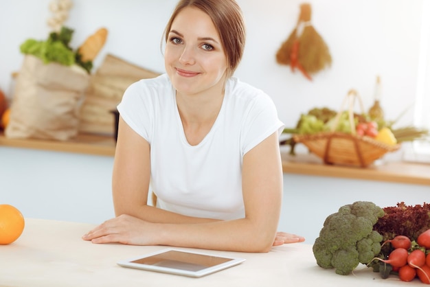 Una joven atractiva que busca una nueva receta para una deliciosa mezcla de ensaladas mientras se sienta a la mesa en la cocina soleada Tablet pc es el mejor libro de cocina