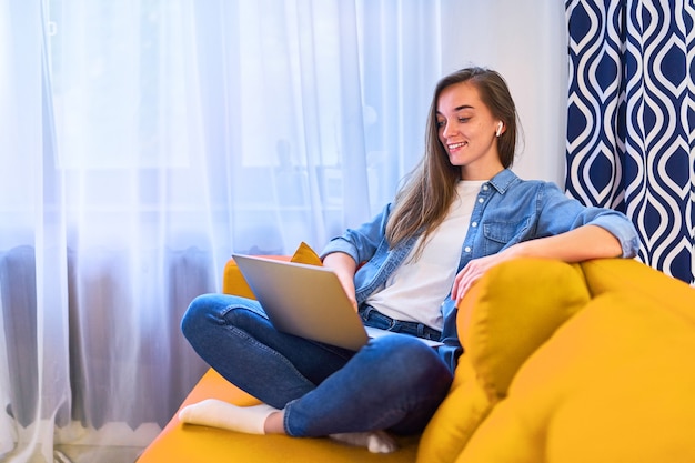 Joven atractiva sonriente feliz linda milenaria sentada en el sofá usando una computadora portátil y auriculares inalámbricos para ver video seminarios web en línea, videollamadas y aprender cursos de idiomas en casa