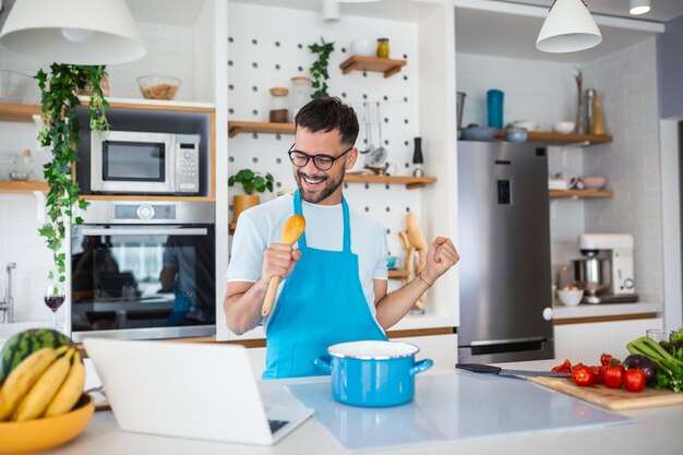 Joven atractivo bailando y cantando en la cocina foto de stock hombre feliz sosteniendo utensilios de cocina como micrófono escuchando música bailando haciendo tareas domésticas en casa preparando el desayuno