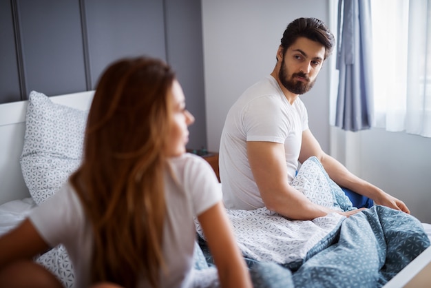 Joven atractivo barbudo mirando a su infeliz novia o esposa mientras está sentado al otro lado de la cama en casa o en el hotel.