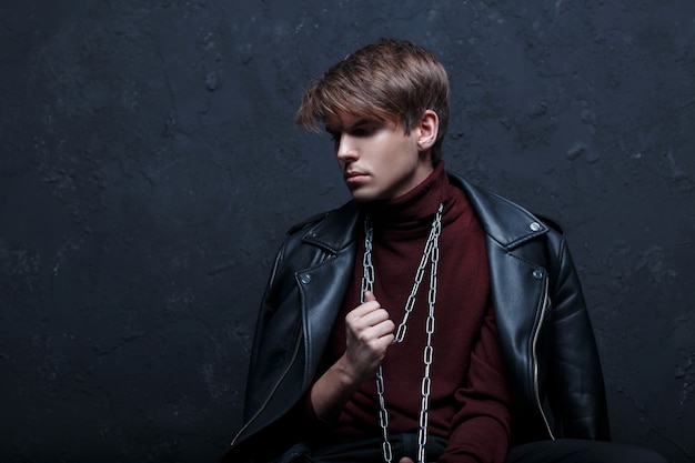 Un joven atractivo con una chaqueta de cuero en un cálido color burdeos golf con una cadena de plata metálica alrededor del cuello, posando en un estudio oscuro contra una pared negra. Chico lindo de la moda