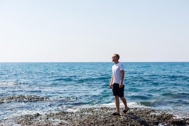 Joven atractivo con gafas de sol en una camiseta blanca y pantalones cortos se encuentra en la orilla del mar mediterráneo