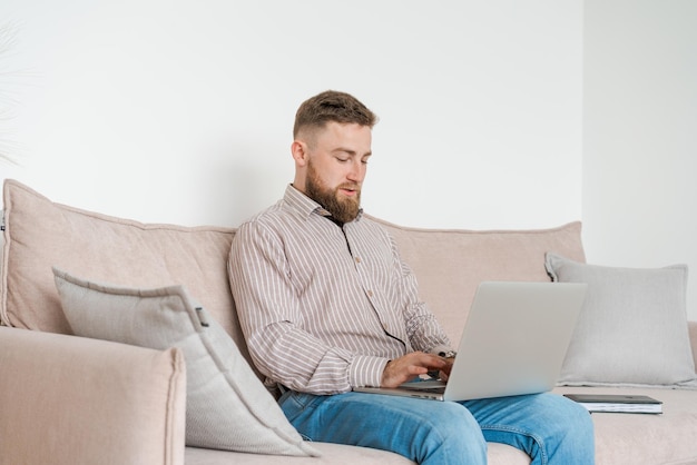 Joven atractivo hombre barbudo sonriente navegando en su computadora portátil mientras está sentado en casa