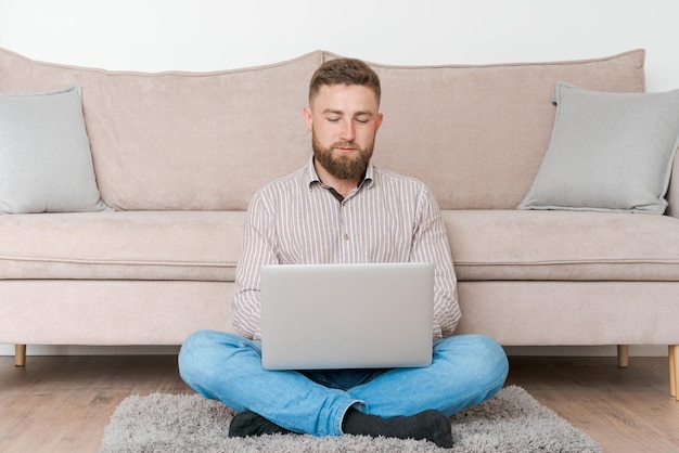Joven atractivo hombre barbudo sonriente navegando en su computadora portátil mientras está sentado en casa
