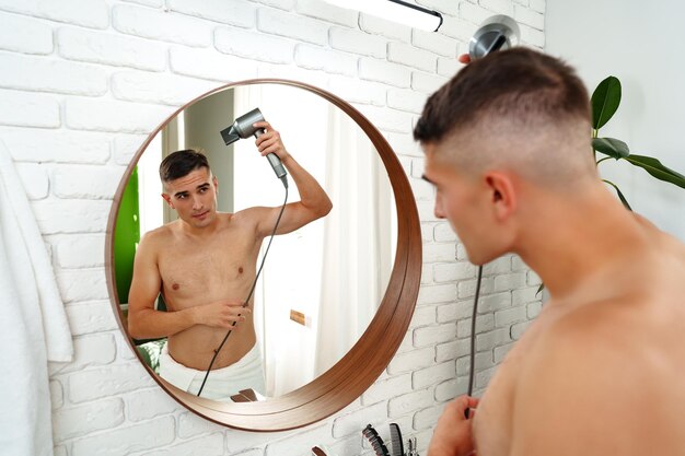 Joven atractivo secándose el cabello después de la ducha en el baño