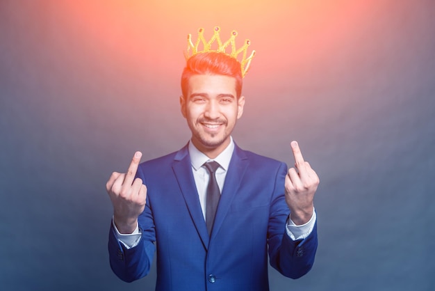 Joven atractivo con un traje azul con una corona en la cabeza que muestra los dedos medios en el resplandor brillante