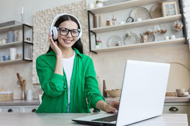 Una joven con auriculares se sienta en casa en la cocina frente a una computadora portátil y graba un podcast