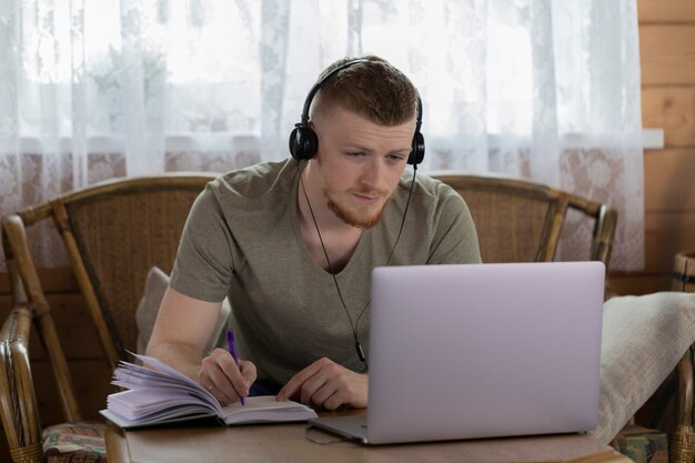 Joven en auriculares trabaja con laptop haciendo notas al diario en casa de madera