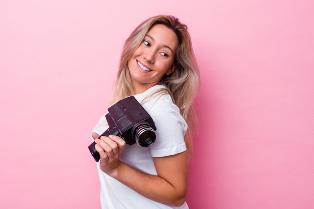 Foto joven australiana filmando con una cámara de video vintage aislada se ve a un lado sonriente, alegre y agradable.