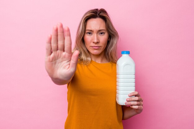 Joven australiana sosteniendo una botella de leche aislada sobre fondo rosa de pie con la mano extendida mostrando la señal de stop, impidiéndote.