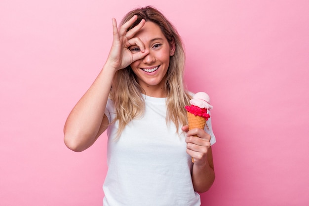Joven australiana sosteniendo un helado aislado sobre fondo rosa emocionado manteniendo el gesto ok en el ojo.