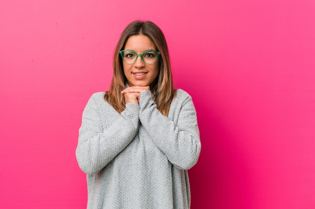 La joven y auténtica y carismática mujer real contra una pared mantiene las manos debajo de la barbilla y mira felizmente a un lado.