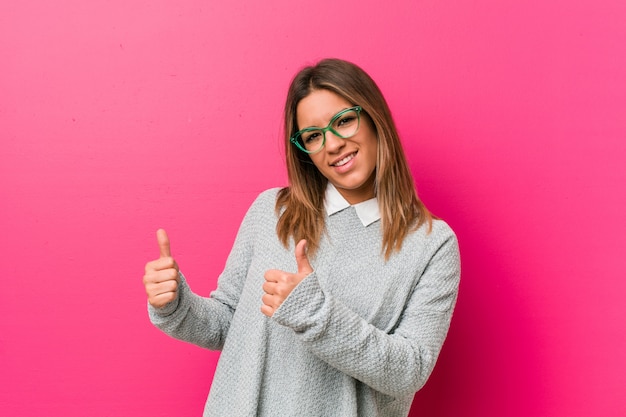 Joven auténtica mujer carismática de personas reales en la pared levantando ambos pulgares, sonriente y confiado.