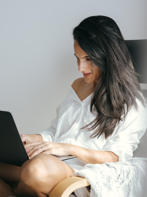 Una joven autónoma étnica concentrada con el pelo largo y oscuro y una camisa blanca sentada en una silla y escribiendo en una computadora portátil mientras trabaja en línea en una computadora portátil en casa