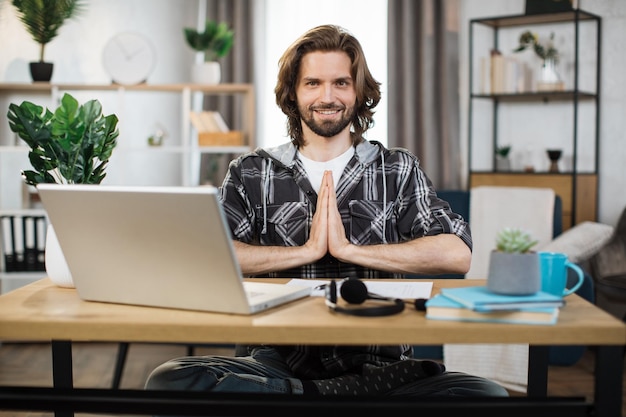 Joven autónomo barbudo sentado en la mesa con los ojos abiertos y aliviando el estrés mediante la meditación