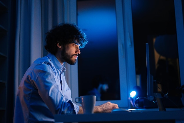 Un joven autónomo exhausto mirando en el monitor, trabajando escribiendo en el teclado y bebiendo café caliente de una taza sentado en el escritorio de la oficina en casa en una habitación oscura. Concepto de trabajo remoto.