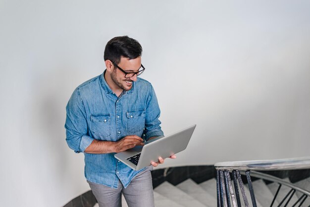 Joven autónomo sonriente trabajando en una laptop mientras sube escaleras en casa