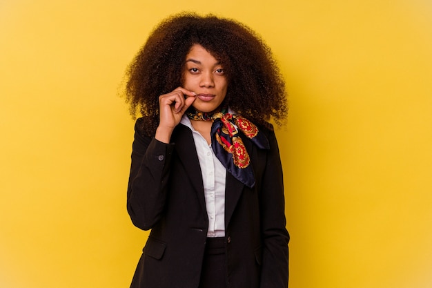 Joven azafata afroamericana aislada en amarillo con los dedos en los labios guardando un secreto.