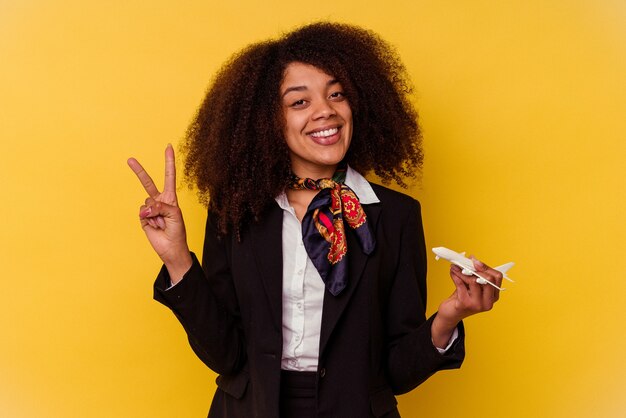 Joven azafata afroamericana sosteniendo un pequeño avión aislado sobre fondo amarillo que muestra el número dos con los dedos.