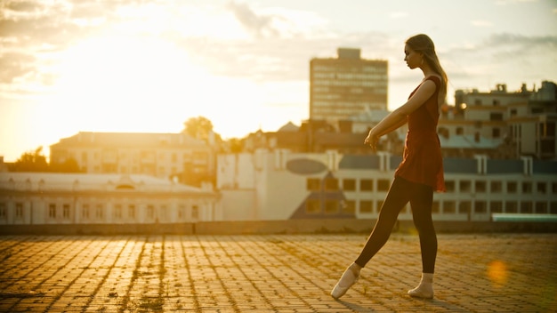 Joven bailarina agraciada de pie en la pose en el techo puesta de sol brillante