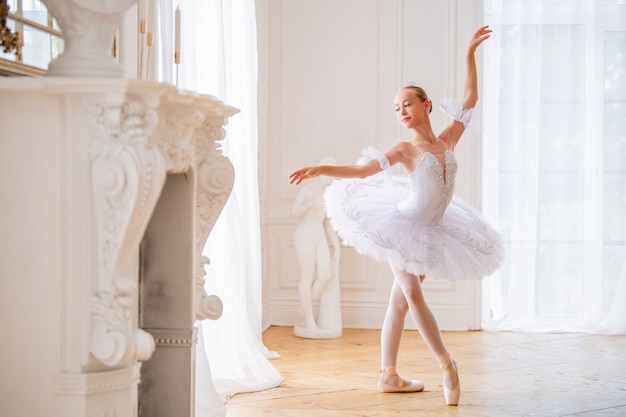 Joven bailarina delgada en un tutú blanco con zapatos de punta está bailando en un gran salón blanco hermoso.