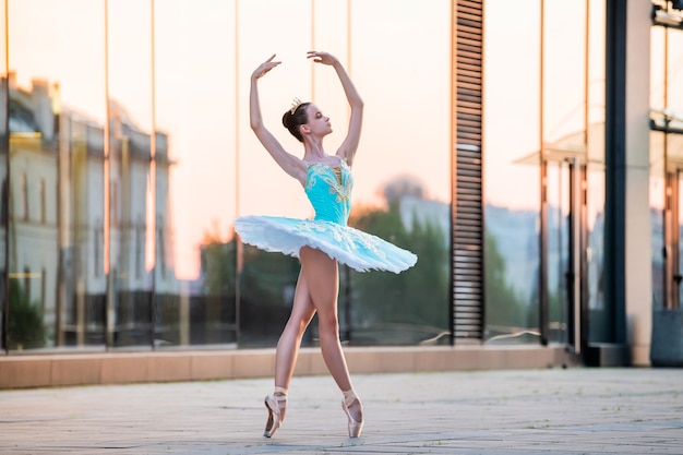 Joven bailarina está bailando en zapatillas de punta en un delicado tutú azul menta con el telón de fondo del reflejo de una puesta de sol en la ciudad