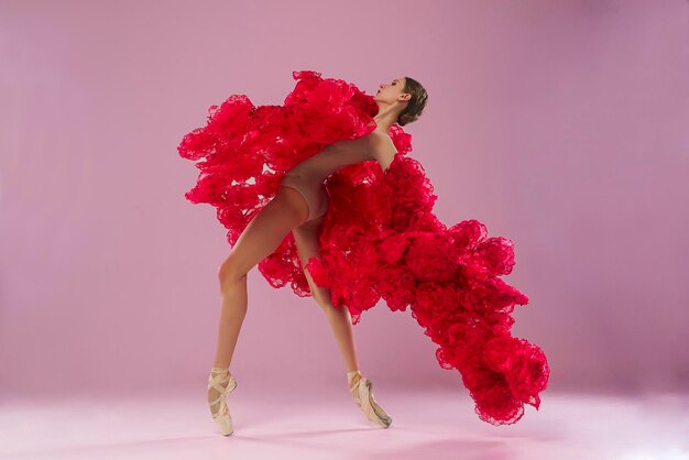 una joven bailarina en un estudio fotográfico en un vestido de capa hecho de flores de rosa muestra movimientos de ballet