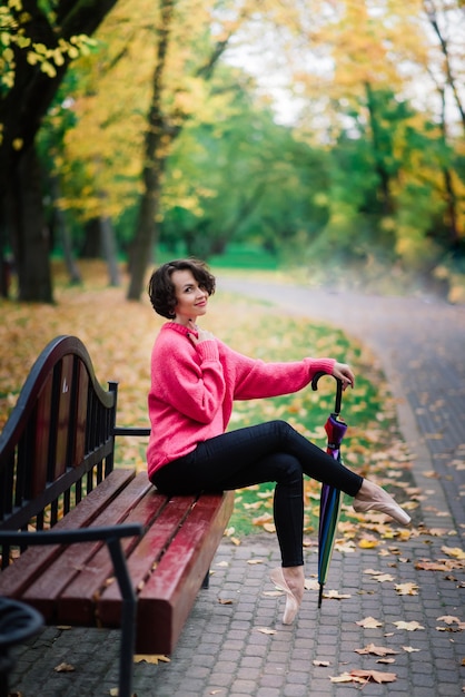 Joven bailarina hermosa en abrigo justo y zapatos de punta se sienta en el banco, descansa al aire libre en el parque de otoño.