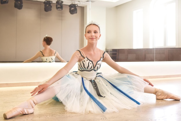 Una joven bailarina practicando poses de ballet sentada contra el espejo en el estudio de baile