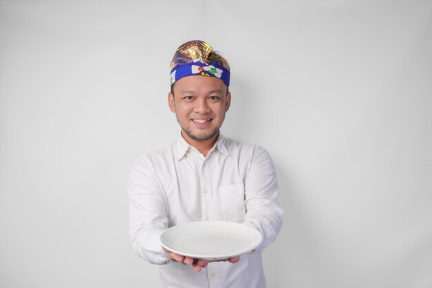 Un joven balinés sonriente con camisa blanca y tocado tradicional mostrando y presentando un plato vacío con espacio para copiar