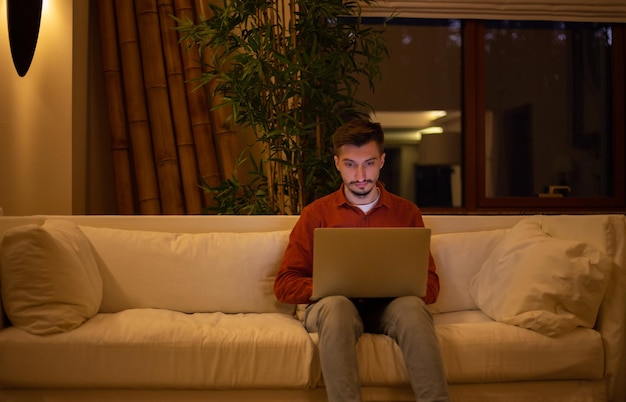 Un joven con barba y camisa roja trabaja con una computadora portátil y se sienta en el sofá por la noche en la casa.