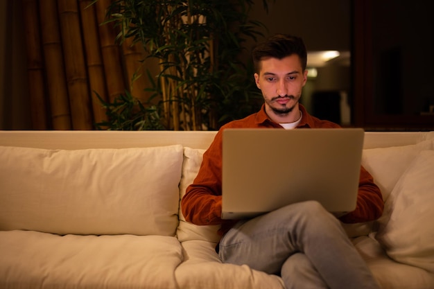 Un joven con barba y camisa roja trabaja con una computadora portátil y se sienta en el sofá por la noche en la casa.