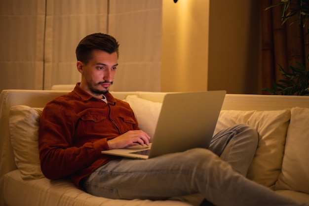 Un joven con barba y camisa roja trabaja con una computadora portátil y se sienta en el sofá por la noche en la casa.