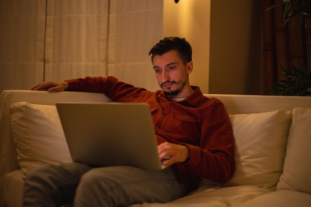 Un joven con barba y camisa roja trabaja con una computadora portátil y se sienta en el sofá por la noche en la casa.