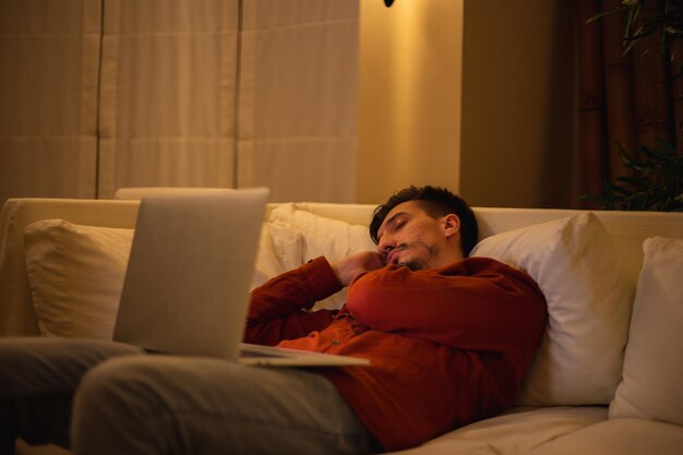 Un joven con barba y camisa roja trabaja con una computadora portátil y se sienta en el sofá por la noche en la casa.