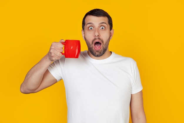 Joven con barba y camiseta blanca bebe una taza de café, asustado en estado de shock con una cara de sorpresa, asustado y emocionado con una expresión de miedo.