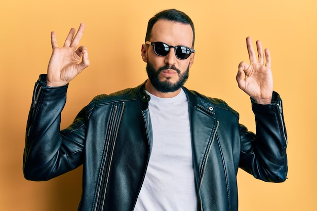 Un joven con barba con chaqueta de cuero negro y gafas de sol se relaja y sonríe con los ojos cerrados haciendo gestos de meditación con los dedos. concepto de yoga