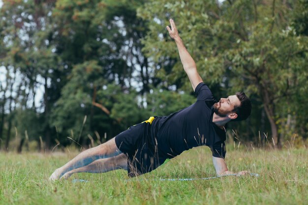 Un joven con barba está haciendo fitness en el parque, realizando ejercicios sobre una colchoneta en el césped